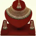 Premium matte gold bridal necklace set with ruby and green stones, heavy choker design, hanging green beads, matching earrings and maang tikka displayed on red stand, Ishwar Arts and Crafts