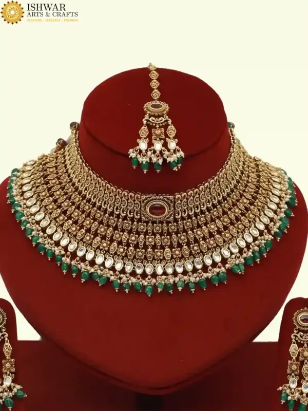 Premium matte gold bridal necklace set with ruby and green stones, heavy choker design, hanging green beads, matching earrings and maang tikka displayed on red stand, Ishwar Arts and Crafts