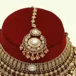 Heavy matte gold brass bridal necklace set with kundan stones, earrings, and maang tikka displayed on red jewelry stand, Ishwar Arts and Crafts