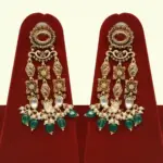 Premium matte gold bridal necklace set with ruby and green stones, heavy choker design, hanging green beads, matching earrings and maang tikka displayed on red stand, Ishwar Arts and Crafts