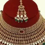 Premium matte gold bridal necklace set with ruby and green stones, heavy choker design, hanging green beads, matching earrings and maang tikka displayed on red stand, Ishwar Arts and Crafts