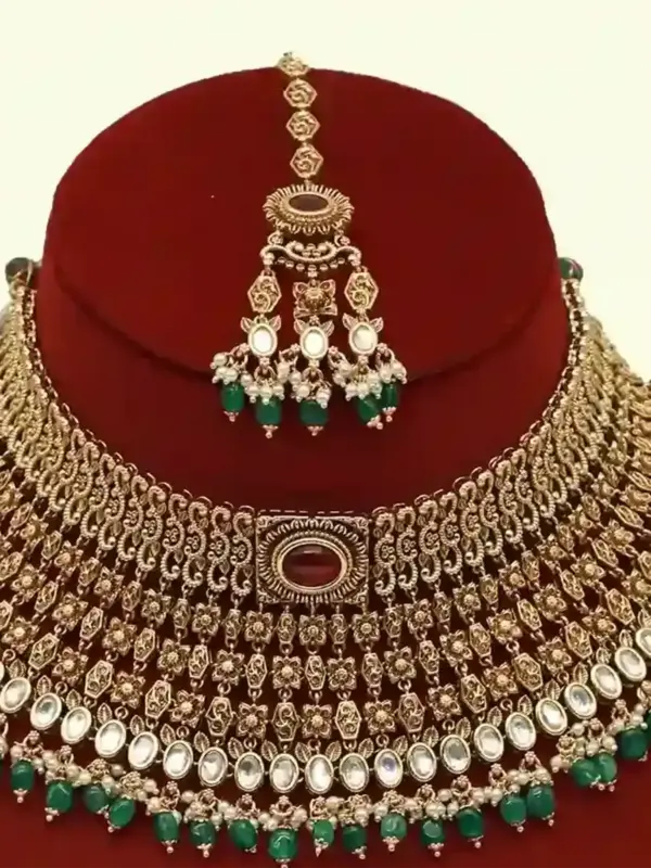 Premium matte gold bridal necklace set with ruby and green stones, heavy choker design, hanging green beads, matching earrings and maang tikka displayed on red stand, Ishwar Arts and Crafts