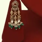 Premium matte gold bridal necklace set with ruby and green stones, heavy choker design, hanging green beads, matching earrings and maang tikka displayed on red stand, Ishwar Arts and Crafts