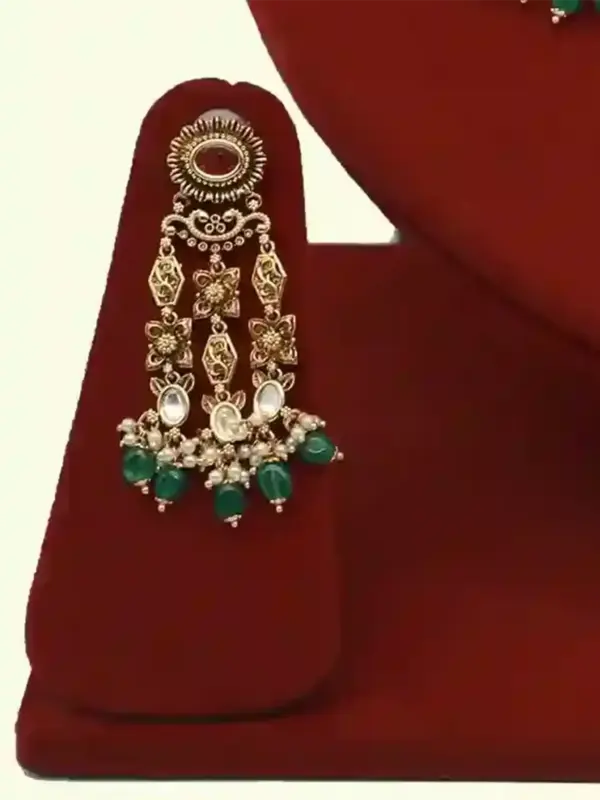 Premium matte gold bridal necklace set with ruby and green stones, heavy choker design, hanging green beads, matching earrings and maang tikka displayed on red stand, Ishwar Arts and Crafts