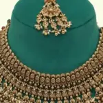 Antique gold intricate carving bridal necklace set with matching heavy earrings and maang tikka displayed on green jewelry stand, Ishwar Arts and Crafts